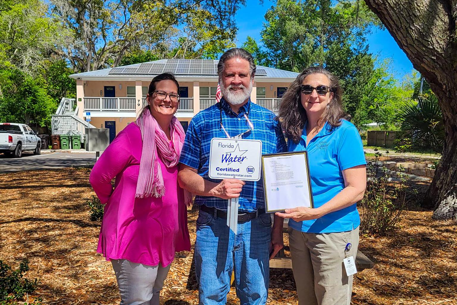 Three people stand infront of a building holding a certificate