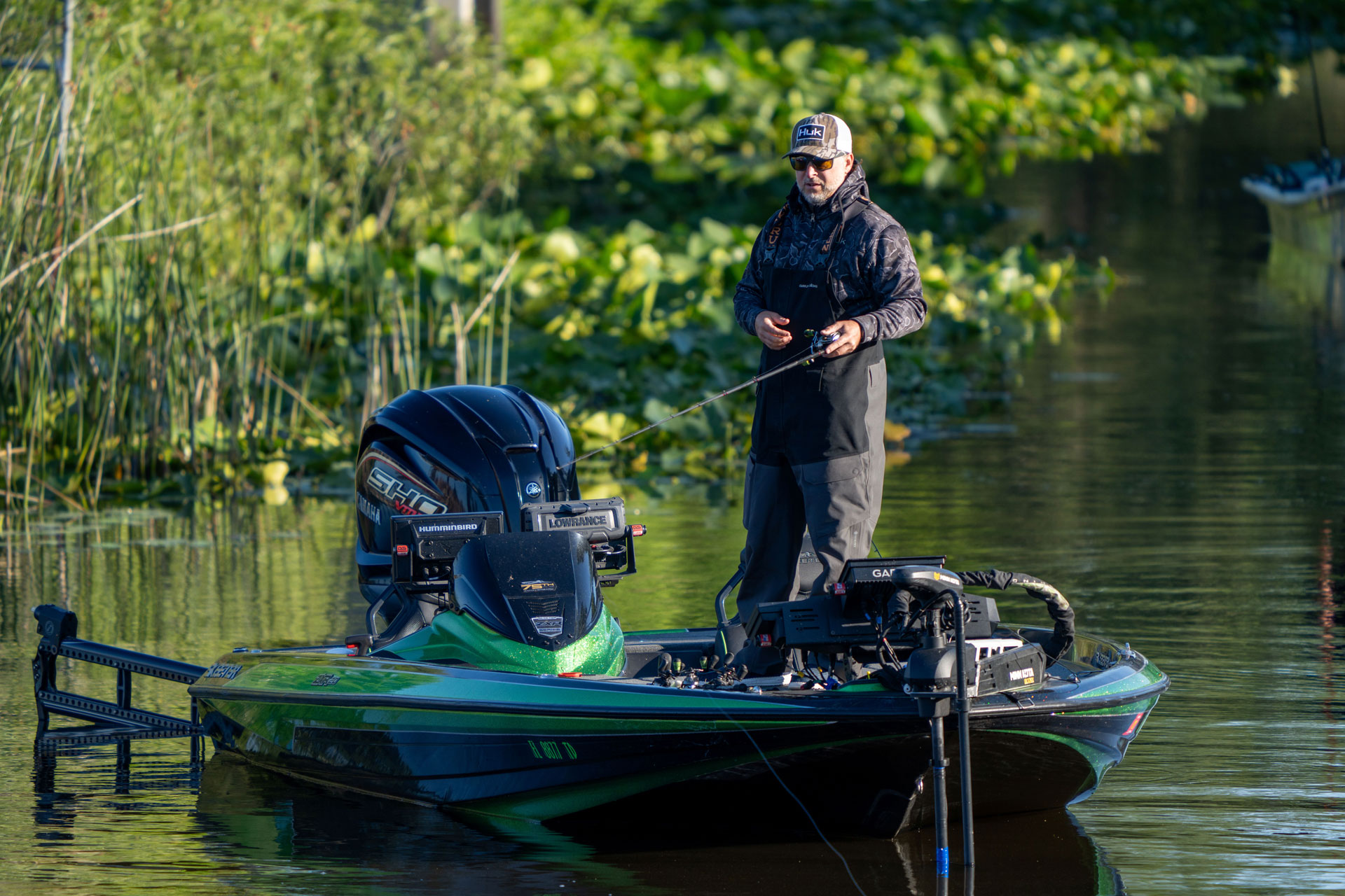 Man standing in boat fishing near shoreline vegetation
