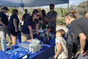St. Johns River Water Management District outreach staff engaging with families at an outdoor educational booth under a blue canopy tent, with children and adults gathered around tables displaying informational materials and activities