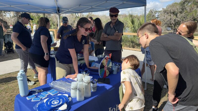St. Johns River Water Management District outreach staff engaging with families at an outdoor educational booth under a blue canopy tent, with children and adults gathered around tables displaying informational materials and activities