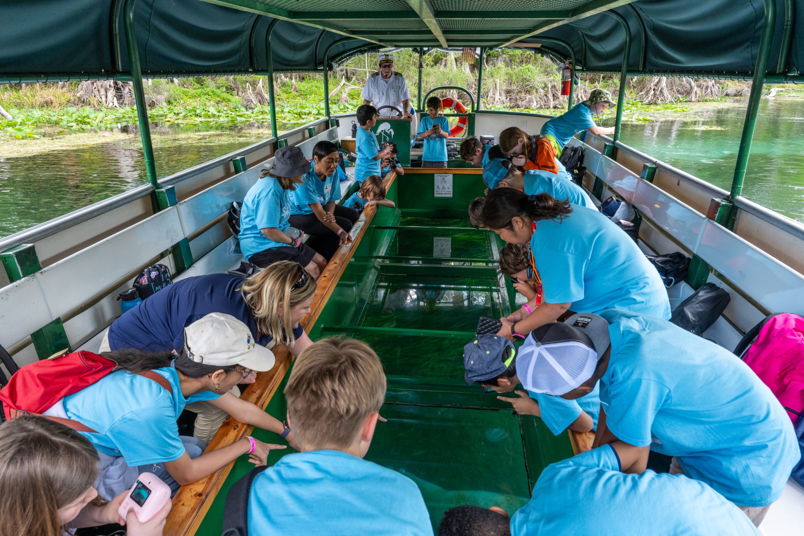 A group of students on a glass‑bottom boat at Silver Springs, looking down through the clear water into the spring basin while the boat glides over the submerged vegetation and limestone bottom.