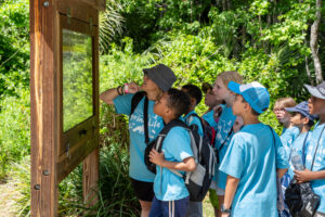 Students in blue “Spring Life” field-trip shirts gather with their teacher at an outdoor kiosk, closely reading an interpretive sign.