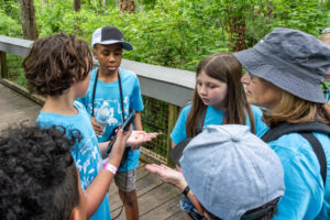 Students in matching blue field-trip shirts gather on a shaded boardwalk, holding out their hands and a smartphone as they examine a small bug together against a backdrop of dense green vegetation.