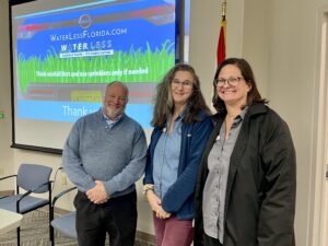 St. Johns River Water Management District water conservation coordinators Kraig McLane, Deirdre Irwin, and Gretchen Smith pose together in front of a projected Water Less Florida campaign slide promoting rainfall-first irrigation practices.