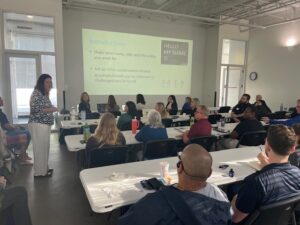 Susan Pokorny of JEA presents an introductory conservation session to a group of attendees seated at white tables, with a projected slide displaying introductory prompts on the screen behind her