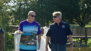Ocklawaha River Strategic Basin Planning Coordinator Jim Peterson and Friends of Lake Apopka Executive Director Joe Dunn walk and converse on a wooden boardwalk surrounded by wetland vegetation and trees.