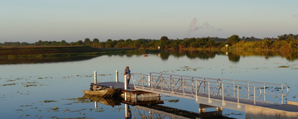 A person fishing from a floating dock with an aluminum access ramp at a calm, vegetated waterway surrounded by Florida wetland vegetation at dusk.