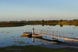 A person fishing from a floating dock with an aluminum access ramp at a calm, vegetated waterway surrounded by Florida wetland vegetation at dusk.