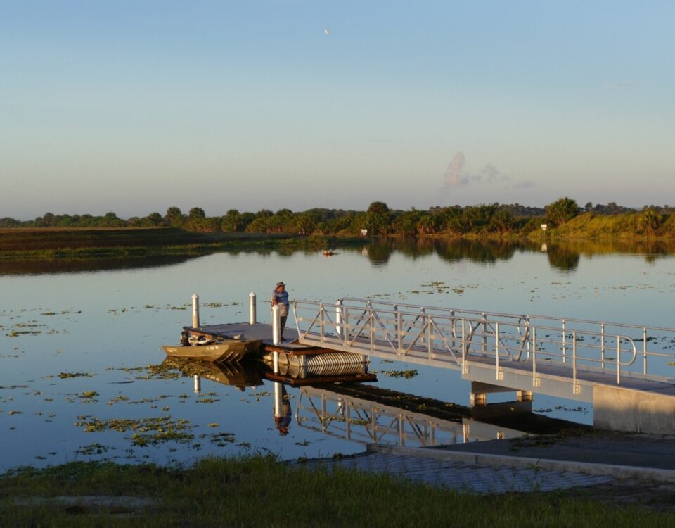 A person fishing from a floating dock with an aluminum access ramp at a calm, vegetated waterway surrounded by Florida wetland vegetation at dusk.