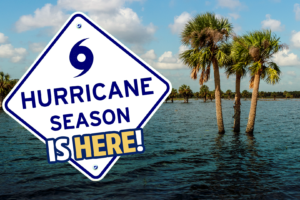 Graphic announcing hurricane season. A large road-sign-style graphic in the foreground displays the words “HURRICANE SEASON IS HERE!” with a hurricane symbol above the text. In the background, several palm trees stand in flooded water beneath a partly cloudy blue sky, illustrating coastal flooding and storm impacts associated with hurricanes.