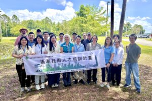 A group of Taiwan delegates and district staff stand together outdoors near a pond, holding a horizontal banner with Chinese text about a water technology training program, with trees, blue sky, and parking area in the background.