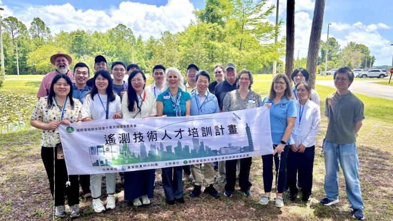 A group of Taiwan delegates and district staff stand together outdoors near a pond, holding a horizontal banner with Chinese text about a water technology training program, with trees, blue sky, and parking area in the background.