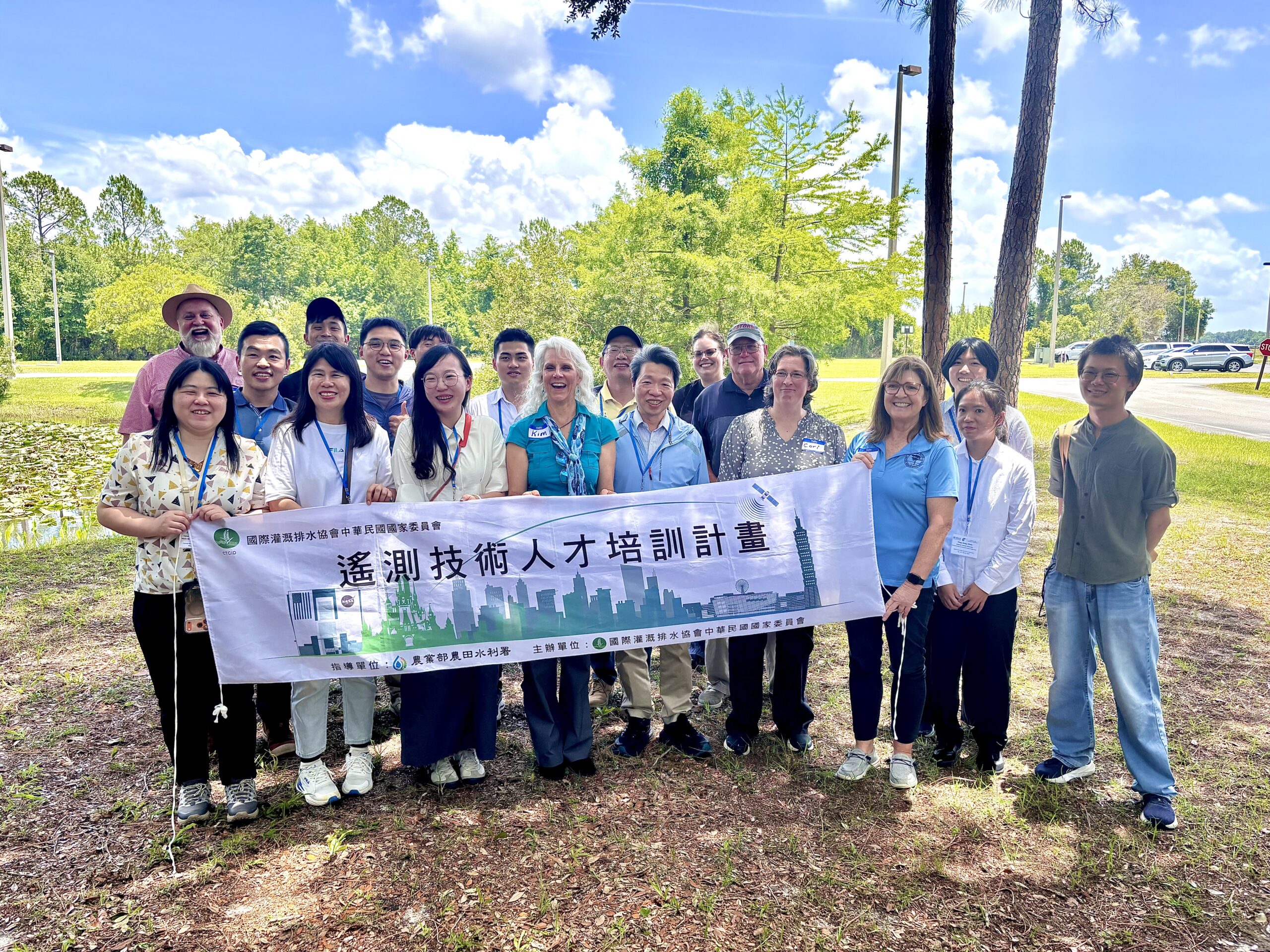 A group of Taiwan delegates and district staff stand together outdoors near a pond, holding a horizontal banner with Chinese text about a water technology training program, with trees, blue sky, and parking area in the background.