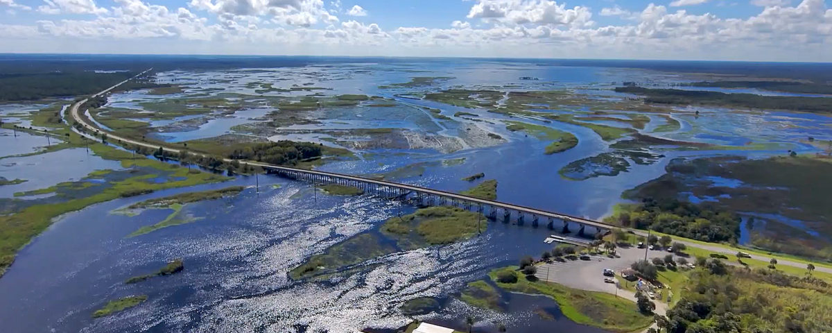 Aerial view of a long highway bridge crossing a wide, shimmering river and marsh system, with patches of open water and wetlands stretching to the horizon under a bright blue, partly cloudy sky, and a small riverside parking and boat access area in the foreground, illustrating regional water management and transportation across the floodplain.