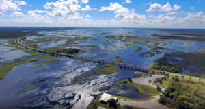 Aerial view of a long highway bridge crossing a wide, shimmering river and marsh system, with patches of open water and wetlands stretching to the horizon under a bright blue, partly cloudy sky, and a small riverside parking and boat access area in the foreground, illustrating regional water management and transportation across the floodplain.