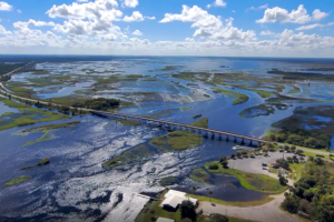 Aerial view of a long highway bridge crossing a wide, shimmering river and marsh system, with patches of open water and wetlands stretching to the horizon under a bright blue, partly cloudy sky, and a small riverside parking and boat access area in the foreground, illustrating regional water management and transportation across the floodplain.