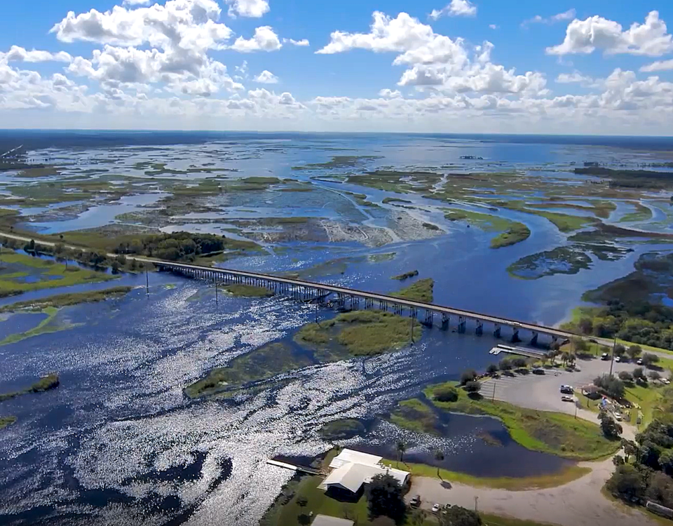 Aerial view of a long highway bridge crossing a wide, shimmering river and marsh system, with patches of open water and wetlands stretching to the horizon under a bright blue, partly cloudy sky, and a small riverside parking and boat access area in the foreground, illustrating regional water management and transportation across the floodplain.