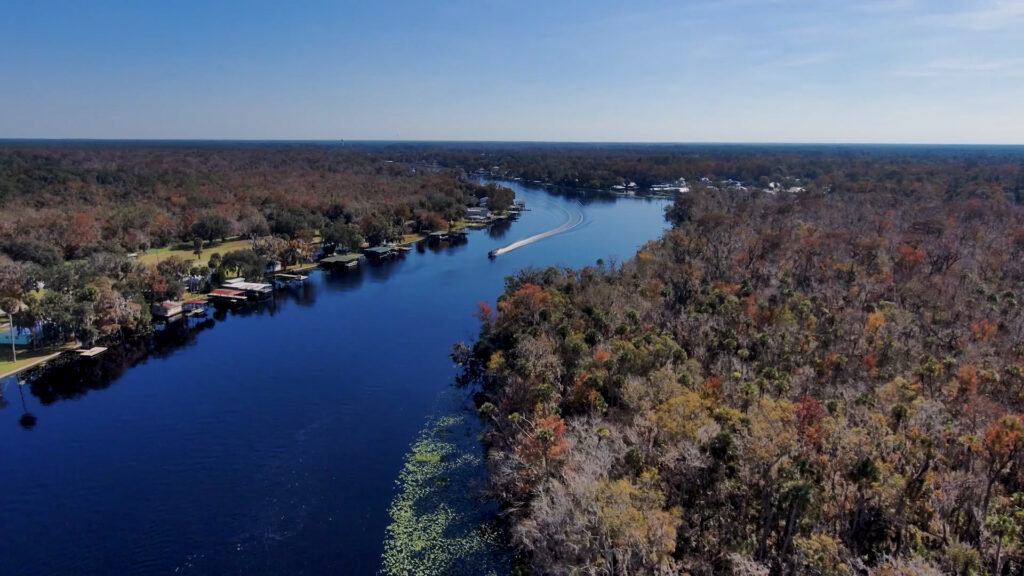 Aerial view of a wide, calm river at Lake George with a motorboat leaving a white wake, bordered by forested shoreline and scattered waterfront homes, illustrating recreational use and natural habitat within the water management district.