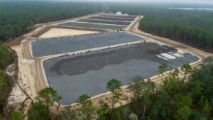Aerial view of a large water management site with multiple lined basins under construction, surrounded by sandy work roads and pine forest, used for storing and treating stormwater before it returns to nearby creeks and wetlands.