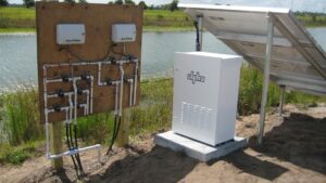 Solar-powered irrigation control equipment sits beside a farm pond, with a wooden panel of valves and pipes connected to a white controller cabinet and solar array used to manage water delivery to nearby fields.
