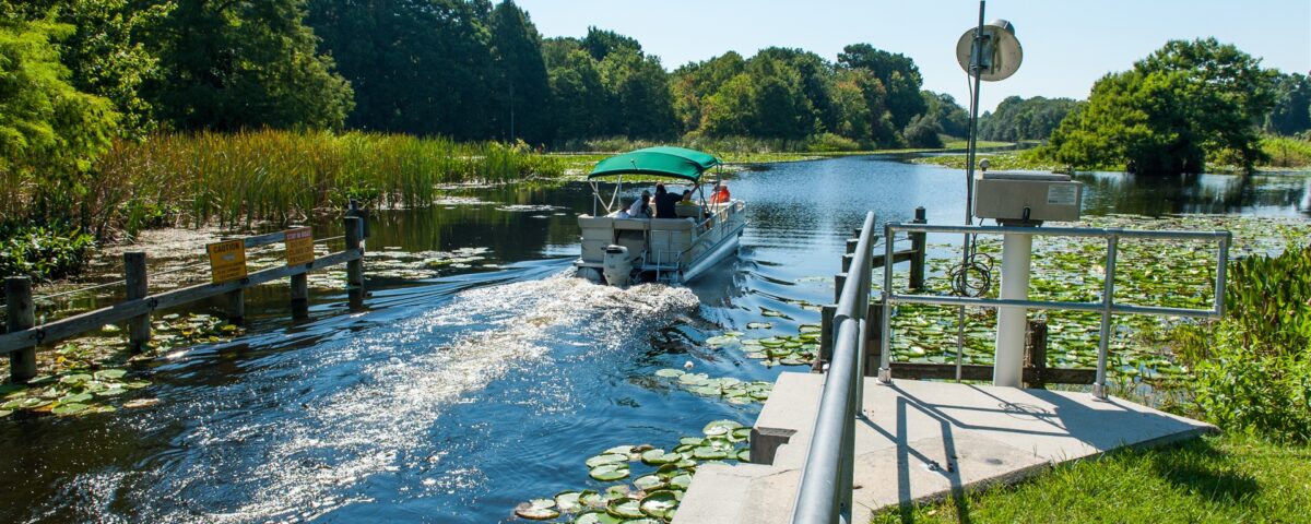Pontoon boat moving through Burrell Lock in the Upper Ocklawaha River Basin, passing lily pads, cattails, warning signs, and monitoring equipment along the managed canal, showing navigable waterway access within the water management district.