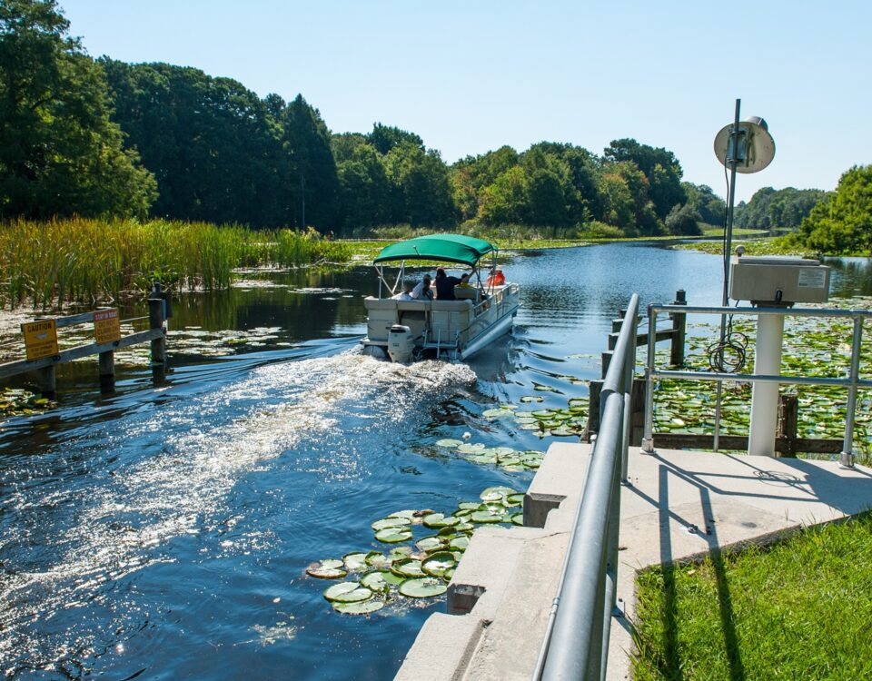 Pontoon boat moving through Burrell Lock in the Upper Ocklawaha River Basin, passing lily pads, cattails, warning signs, and monitoring equipment along the managed canal, showing navigable waterway access within the water management district.
