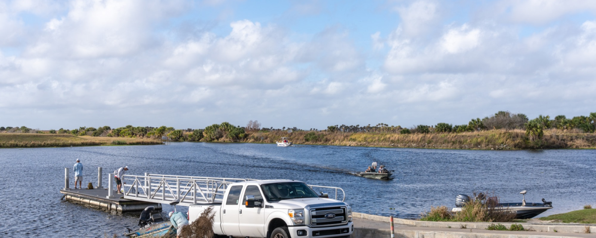 Boat ramp on a river with a white pickup truck, several boats launching and traveling on the water under a partly cloudy sky.