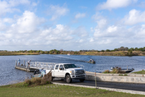 Boat ramp on a river with a white pickup truck, several boats launching and traveling on the water under a partly cloudy sky.