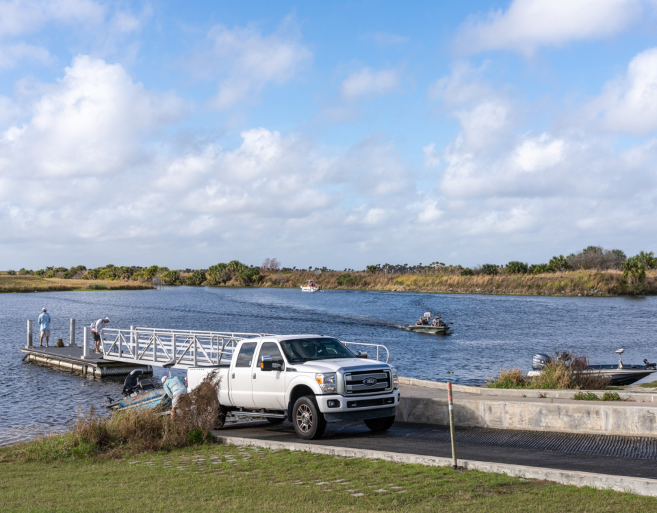 Boat ramp on a river with a white pickup truck, several boats launching and traveling on the water under a partly cloudy sky.