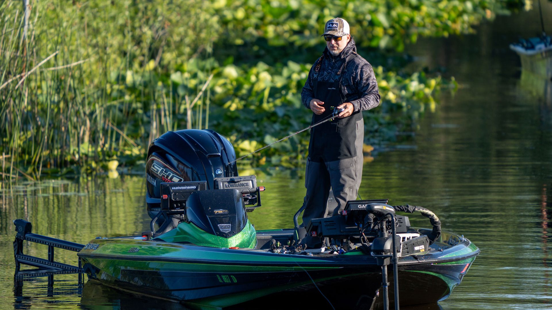 Man standing in boat fishing near shoreline vegetation