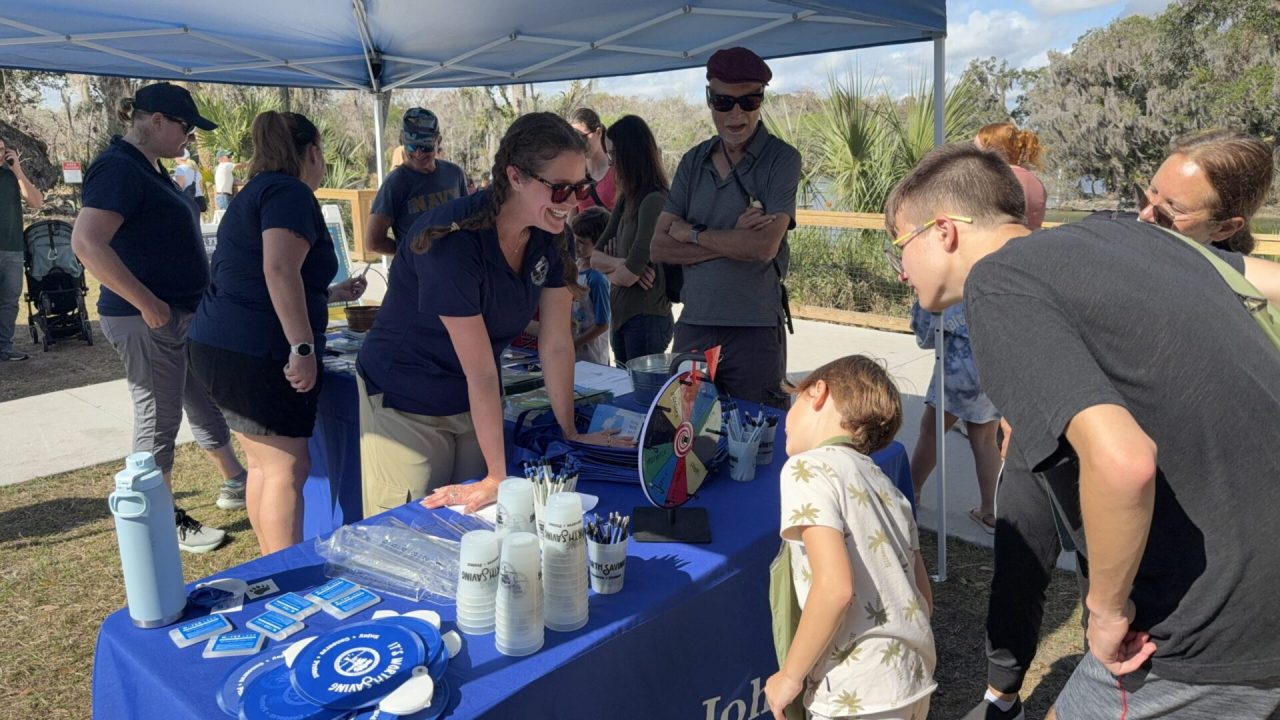 St. Johns River Water Management District outreach staff engaging with families at an outdoor educational booth under a blue canopy tent, with children and adults gathered around tables displaying informational materials and activities