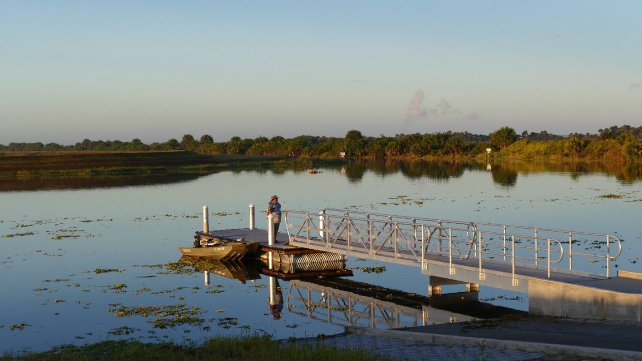 A person fishing from a floating dock with an aluminum access ramp at a calm, vegetated waterway surrounded by Florida wetland vegetation at dusk.