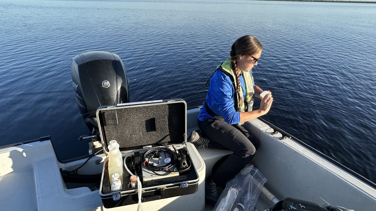 A field technician wearing a life vest examines a water sample on a boat, with field equipment and sample bottles nearby on calm open water.