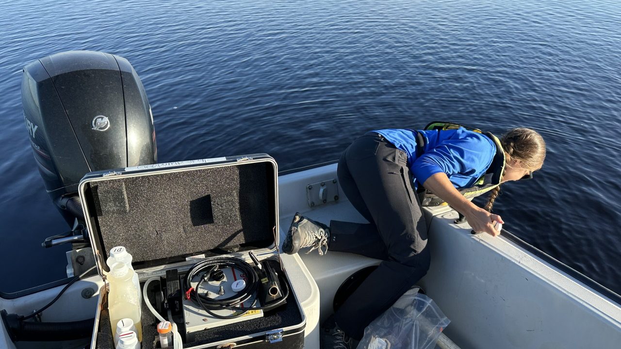 A field technician leans over the side of a boat to collect a water sample, with an open field equipment case and sampling supplies visible on deck.