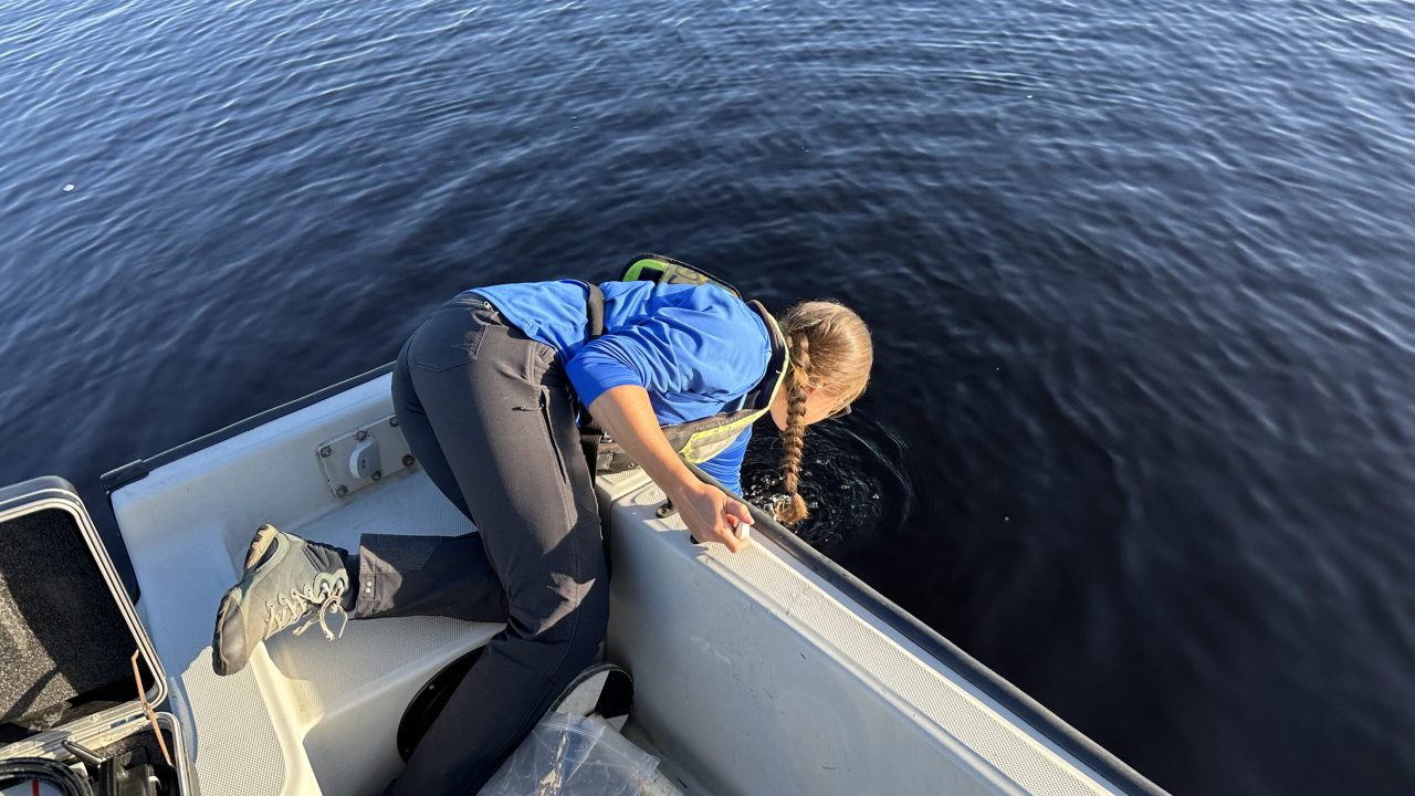 A field technician leans over the side of a boat to collect a water sample, with an open field equipment case and sampling supplies visible on deck.