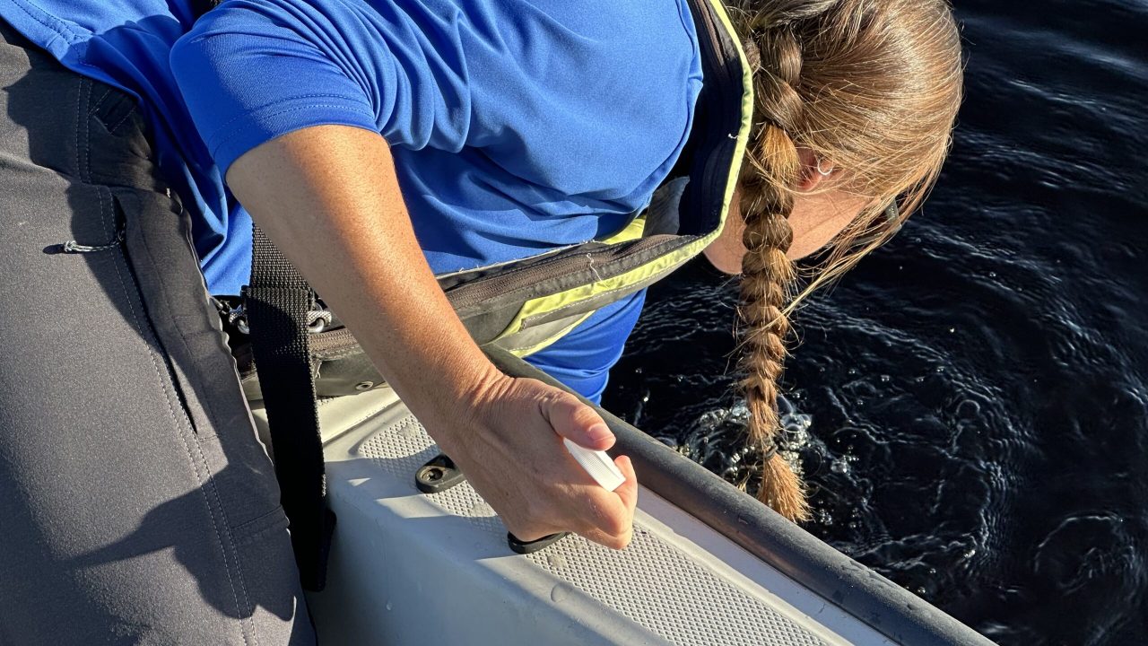 Close-up of a field technician in a life vest leaning over the side of a boat to submerge a sample collection vial into open water.
