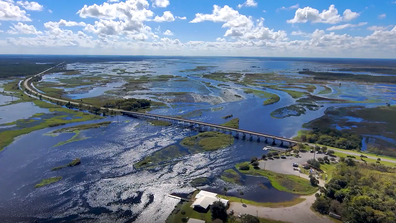 Aerial view of a long highway bridge crossing a wide, shimmering river and marsh system, with patches of open water and wetlands stretching to the horizon under a bright blue, partly cloudy sky, and a small riverside parking and boat access area in the foreground, illustrating regional water management and transportation across the floodplain.