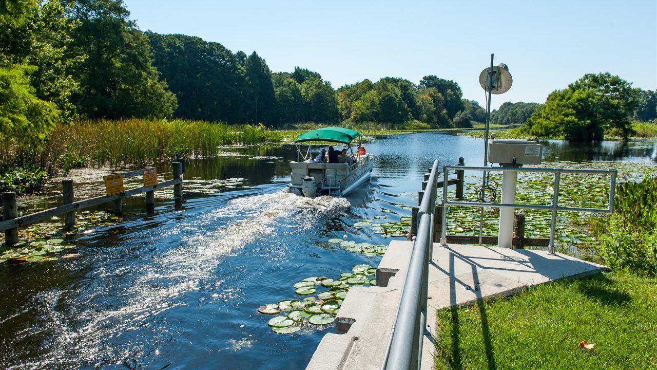Pontoon boat moving through Burrell Lock in the Upper Ocklawaha River Basin, passing lily pads, cattails, warning signs, and monitoring equipment along the managed canal, showing navigable waterway access within the water management district.