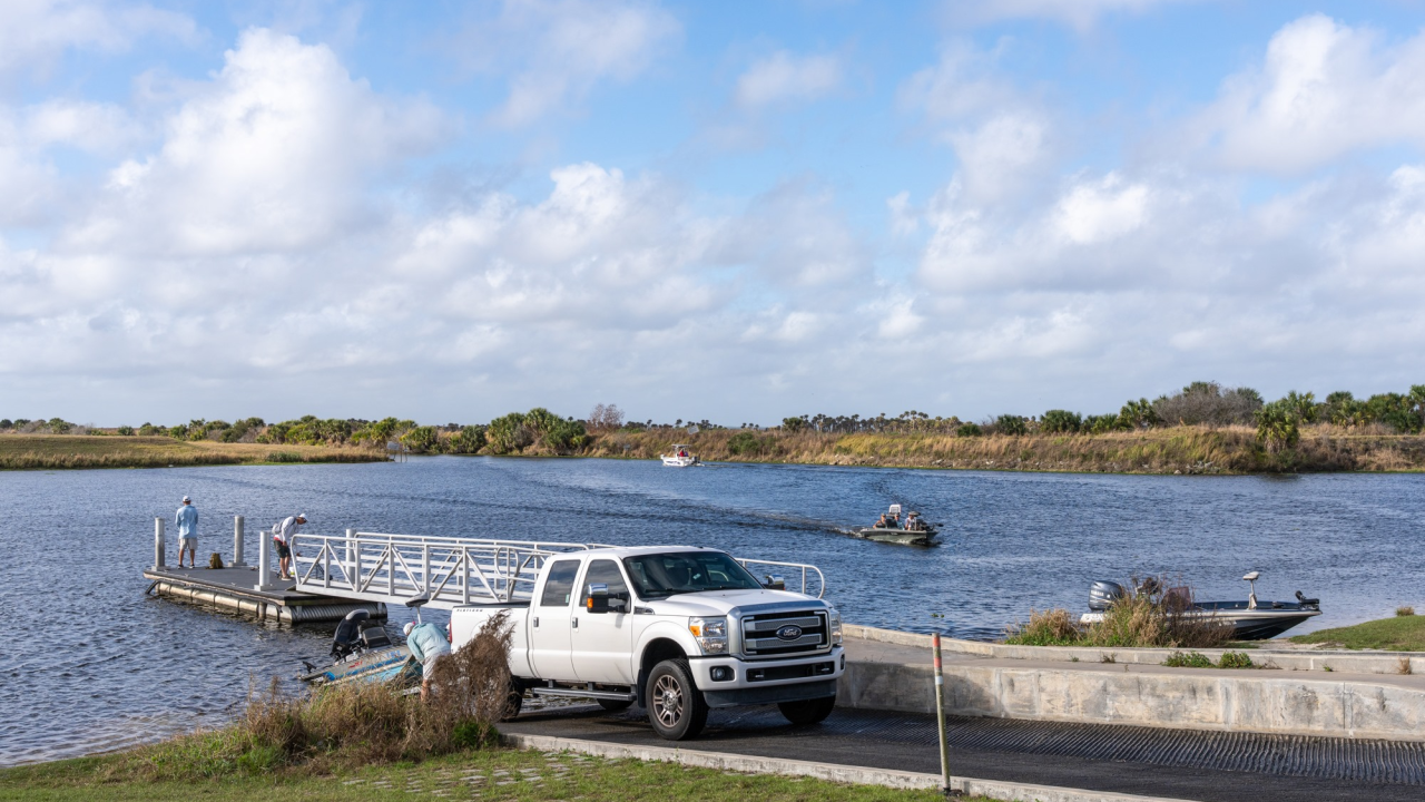 Boat ramp on a river with a white pickup truck, several boats launching and traveling on the water under a partly cloudy sky.